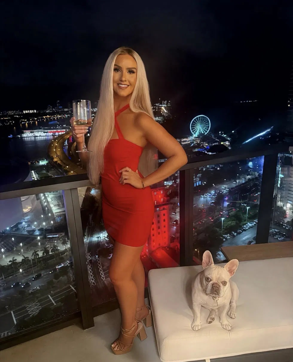 Christine in a red dress with a glass of wine on the balcony at night, Luna sitting beside her, downtown Miami lit up behind